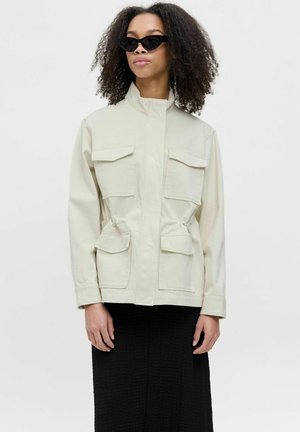 Chaqueta utilitaria de color beige claro con cuatro bolsillos frontales, cintura elástica y cuello. Usada sobre un vestido negro texturizado. Gafas de sol negras completan el look.