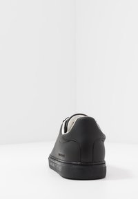 Baskets noires avec un dessus en cuir lisse, accents blancs sur le col et une semelle en caoutchouc texturée. Logo visible au niveau du talon.