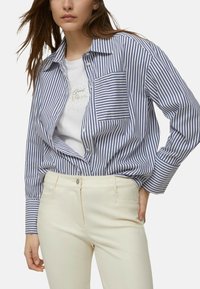Camicia a righe blu e bianca con bottoni e taschino. Indossata sopra una maglietta bianca con dettaglio testuale scintillante. Pantaloni aderenti color crema.