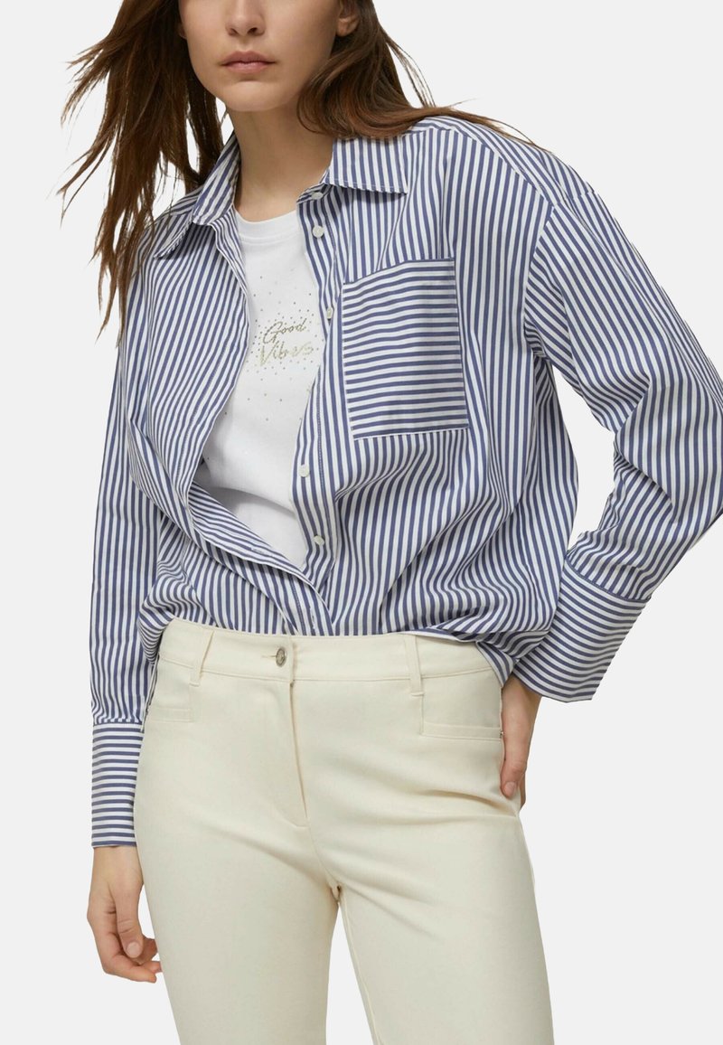 Camicia a righe blu e bianca con bottoni e taschino. Indossata sopra una maglietta bianca con dettaglio testuale scintillante. Pantaloni aderenti color crema.