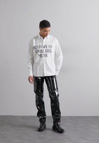 White button-up shirt with "HISTORY OF UNHEARD MUSIC" in black text, paired with shiny black vinyl pants and black ankle boots.
