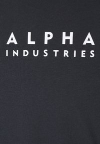 Juodas audinys su ryškiu baltu tekstu "ALPHA INDUSTRIES", su lygiu paviršiumi ir šonkaulinio dizaino.