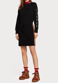 Zwart sweatshirtjurk met lange mouwen, trekkoord in de taille en oranje zijstrepen. Gedragen met rode sokken en bruin veterlaarzen.