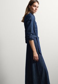 Vestido de mezclilla con mangas largas, cintura anudada y un corte ajustado. Presenta un color azul profundo y un frente de botones con un bolsillo.