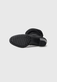 Black suede boot lying on its side showing textured rubber sole and low block heel with brand logo on heel tip.