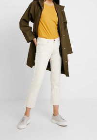 Olivgrön regnjacka, senapsgul t-shirt, vita cropped jeans, ljusgrå sneakers. Har fickor, tryckknappar och råa fållar.