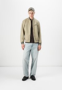 Light beige varsity jacket with black stripes, brown shirt underneath, loose light blue jeans, black shoes, and a gray cap.