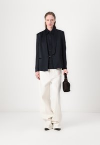 Černý elegantní blejzr přes černou košili, doplněný širokými bílými kalhotami a špičatými černými botami. Malá tmavá taška je držena v ruce.