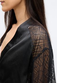 Mustast satiinist hommikumantel keeruka pitsist varrukatega, millel on geomeetrilised mustrid, mis rõhutavad siledat tekstuuri ja õrna detailirohkust.