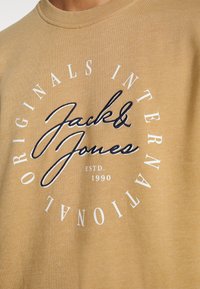 Personne portant un sweat-shirt beige avec "Jack & Jones" brodé en lettres noires cursives et un texte circulaire blanc indiquant "Originals International" et "ESTD. 1990."