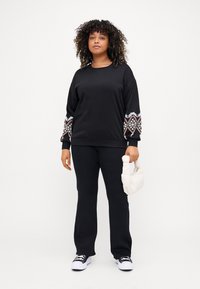 Svart sweatshirt med mönstrade ärmar, tillsammans med svarta byxor och vita sneakers. Håller en liten vit väska. Lockigt hår.