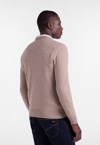 Beige knitted sweater with textured check pattern, ribbed hem and cuffs, worn over dark denim jeans. Model facing away, showcasing back design.