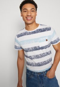 Camiseta blanca de cuello redondo con rayas azules, que presenta un bolsillo en la parte frontal y un pequeño parche de logo marrón. Combinada con jeans de mezclilla azules.