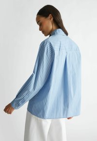 Camisa abotonada de un azul claro con rayas verticales blancas, de mangas largas, con cuello clásico y un pliegue en la parte posterior para facilitar el movimiento.