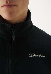 Crna flis jakna s visokim ovratnikom, prednjim patentnim zatvaračem i bijelim "berghaus" logotipom na lijevoj strani prsnog dijela. Mekana tekstura, ležeran dizajn.