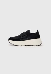 Svart atletisk sneaker med strikket overdel, kontrasterende hvit såle, teksturerte detaljer og en dekorativ spenne. Rund form og lav profil.