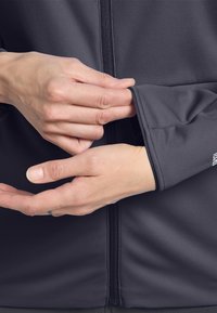 Dettaglio di una giacca sportiva grigia che mostra le mani che aggiustano il polsino. Il tessuto è liscio con una texture morbida e presenta una cerniera frontale.
