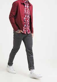 Burgundy shirt jacket over a patterned red and white button-up, paired with grey jeans and white sneakers, featuring a distressed hem.