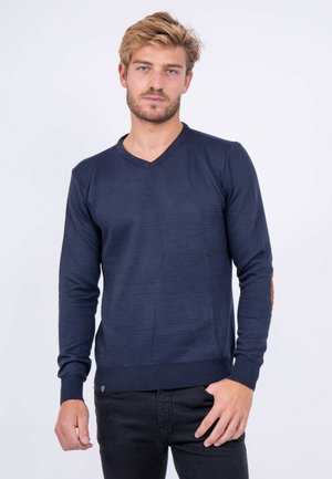 Homme blond avec une légère barbe portant un pull en V bleu marine avec des coudières, debout devant un fond blanc uni.