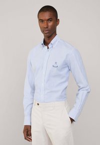Camisa de botones de rayas azules y blancas con mangas largas, que presenta un pequeño logotipo bordado en el pecho y un cuello puntiagudo.