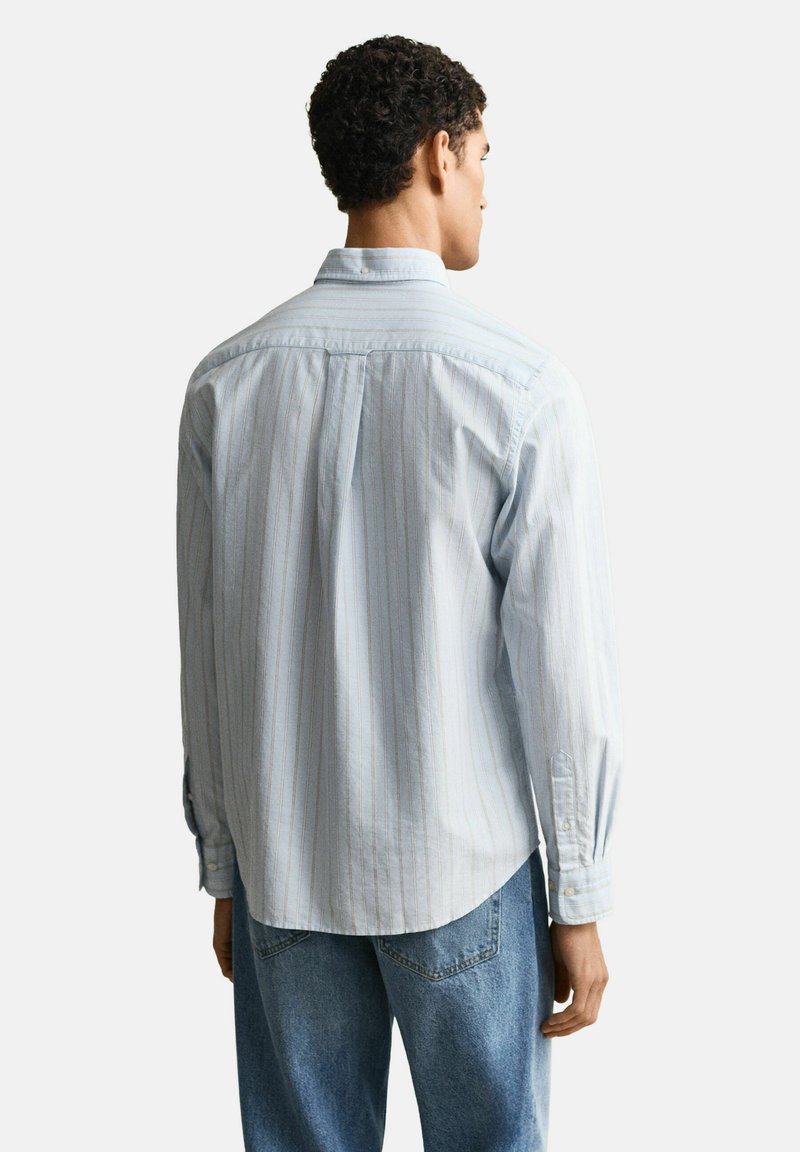 Uomo che indossa una camicia a maniche lunghe a righe azzurro chiaro e jeans blu, in piedi con le spalle rivolte verso l'osservatore su uno sfondo bianco uniforme.