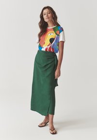 Camiseta gráfica colorida con diseño abstracto combinada con una larga falda envolvente verde. La modelo lleva chanclas marrones. Fondo neutro.