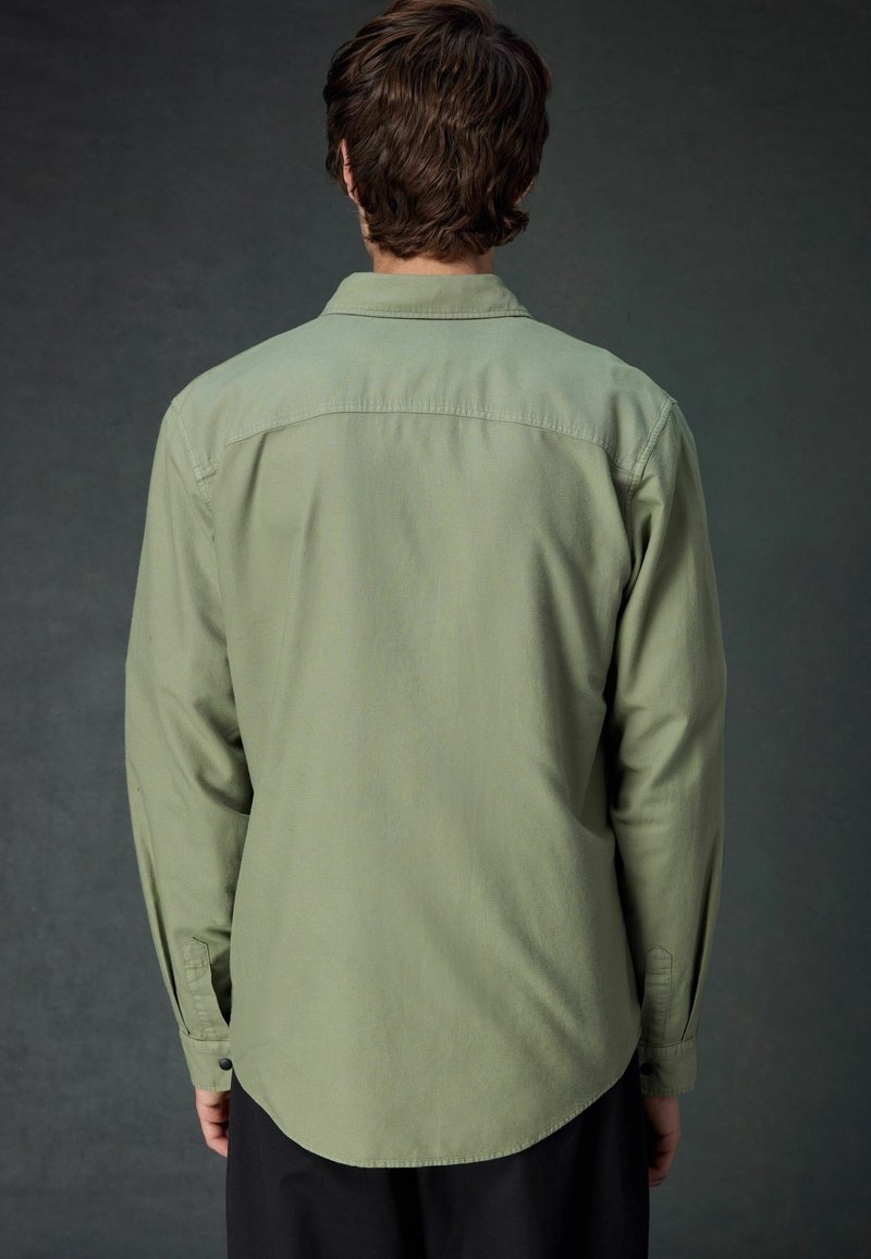 Camicia a maniche lunghe verde oliva con orlo curvo, polsini abbottonati e colletto semplice; texture liscia con una finitura leggermente opaca.