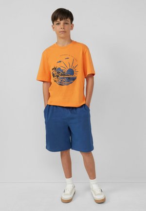 Pomarańczowa koszulka bawełniana z niebieskim graficznym wzorem zachodu słońca i palm, zestawiona z niebieskimi szortami. Sneakersy o białej bazie z beżowymi akcentami.