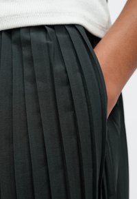 Jupe plissée noire avec une texture fine. Le tissu de la jupe tombe en douceur, mettant en valeur des plis profonds et une poche latérale près de la taille.