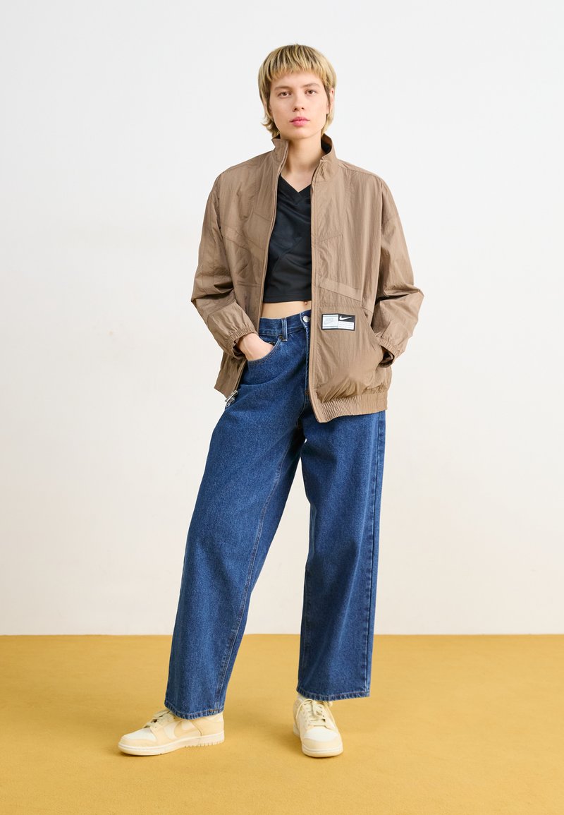 Giacca marrone chiaro con colletto alto, chiusura a zip e etichetta del marchio. Abbinata a jeans a gamba larga blu scuro e sneakers beige su un pavimento giallo.