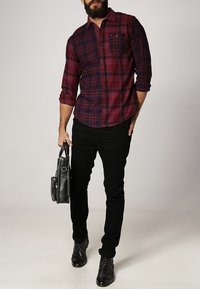 Czerwono-granatowa koszula w kratę z długimi rękawami, połączona z czarnymi spodniami typu slim-fit i eleganckimi czarnymi butami. Trzyma czarną torbę.