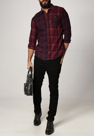 Camisa de cuadros en rojo y azul marino con botones y mangas largas, combinada con pantalones negros ajustados y zapatos negros elegantes. Sosteniendo un bolso negro.