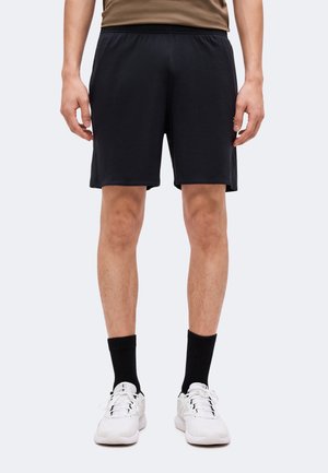 Homme portant un short de sport noir, des chaussettes noires et des baskets blanches, debout, les mains détendues le long du corps.