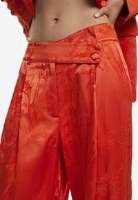 Pantalones de satén rojo con bordado floral, que presentan una parte delantera plana, botones laterales y pliegues; textura suave y corte ajustado.