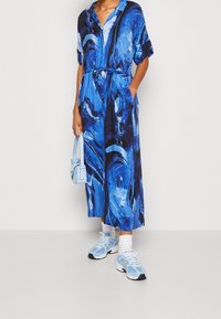 Vestido maxi de mármol azul con un corte relajado, mangas cortas y una cintura con lazo. Combinado con zapatillas ligeras azules y un pequeño bolso.