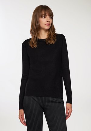 Maglione nero a maniche lunghe in tessuto a maglia, scollatura rotonda, polsini e orlo a costine. Indossato con pantaloni a righe che mostrano una silhouette aderente.