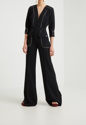 Femme portant une combinaison noire avec un liseré blanc, un décolleté en V profond, des poches à rabat, des manches trois-quarts et un pantalon à jambes larges, debout à l'intérieur.