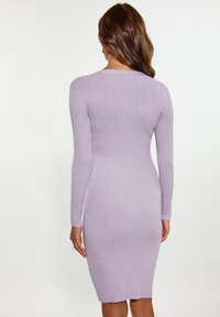 Kvinna med långt brunt hår iförd en figurnära, ribbad, långärmad lavendelfärgad klänning, visad från ryggen mot en enkel vit bakgrund.