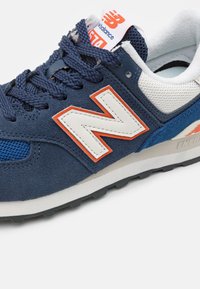 Baskets New Balance bleu marine et blanches avec des accents orange, en mesh et daim, lacets bleu marine, et doublure intérieure rembourrée.