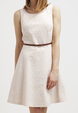Mujer que lleva un vestido sin mangas de color rosa claro con textura, con cuello redondo y un cinturón fino marrón en la cintura.