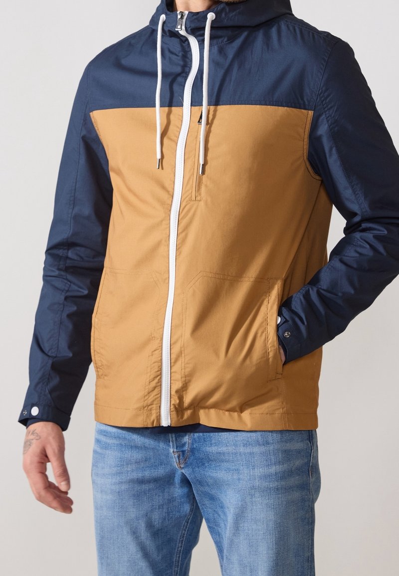 Kleurblokjas met een blauwe capuchon en mouwen, en een tan lichaam. Inclusief een ritssluiting aan de voorkant, een verstelbare capuchon en zijzakken.