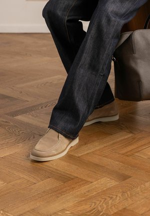 Persoon draagt donkere spijkerbroek en beige suède loafers, houdt een bruine leren tas vast op een houten parketvloer.