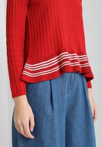 Red knitted sweater with a textured finish, featuring white striped accents at the hem. Paired with loose denim trousers.