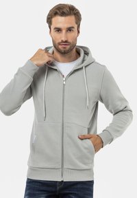 Haut de survêtement zippé gris clair avec capuche à cordon de serrage, poches avant et poignets côtelés. Fabriqué en matériau doux, design minimaliste.