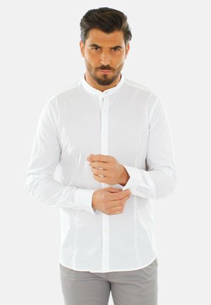 Uomo con capelli scuri che sistema il polsino di una camicia bianca con colletto alla mandarino, indossa pantaloni grigio chiaro, in piedi davanti a uno sfondo uniforme.