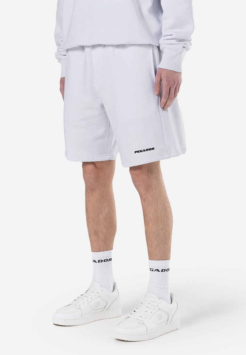Personne portant un short blanc, des chaussettes blanches montantes avec un texte noir, des baskets blanches et un sweat-shirt blanc, debout devant un fond uni.