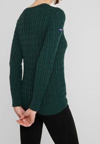 Suéter verde de punto con cable, manga larga, puños acanalados, cuello redondo y un pequeño parche con logo en el brazo izquierdo. La textura es suave y texturizada.