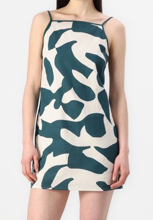 Frau trägt ein ärmelloses Minikleid mit dünnen Trägern und einem abstrakten weißen und dunkelgrünen Muster.