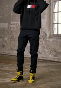 Zwarte hoodie met een rood-wit logo, gecombineerd met zwarte cargobroek en gele hoge sneakers met zwarte accenten.