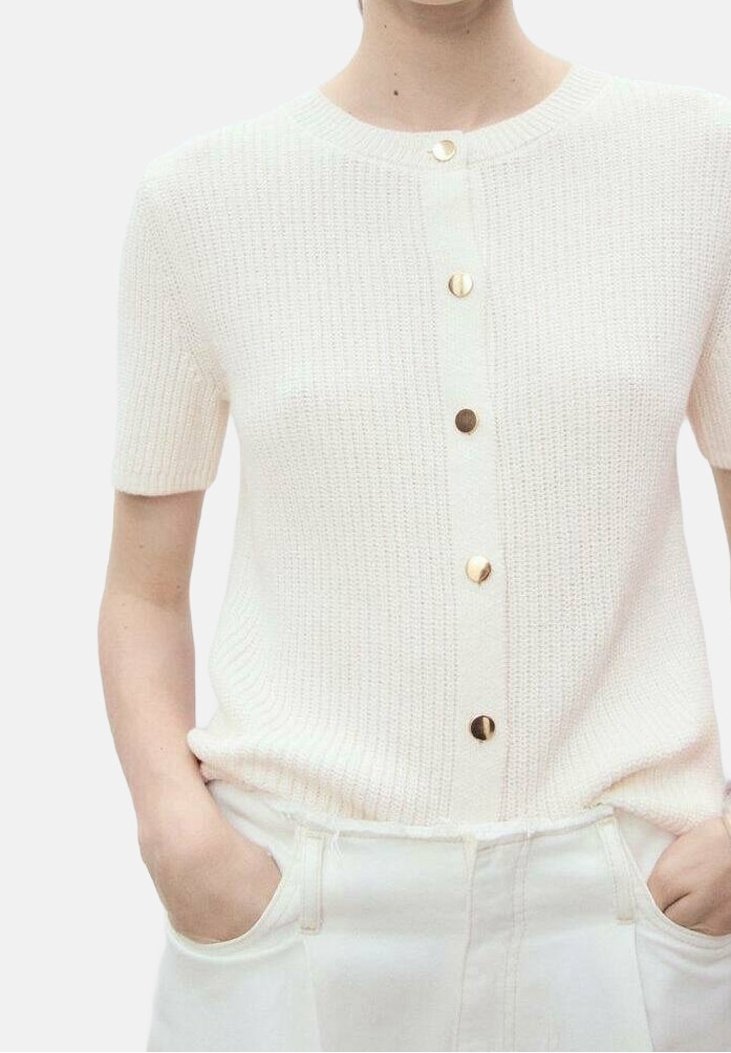 Personne portant un cardigan en maille crème à manches courtes avec des boutons dorés et un pantalon blanc taille haute, les mains dans les poches de manière décontractée.